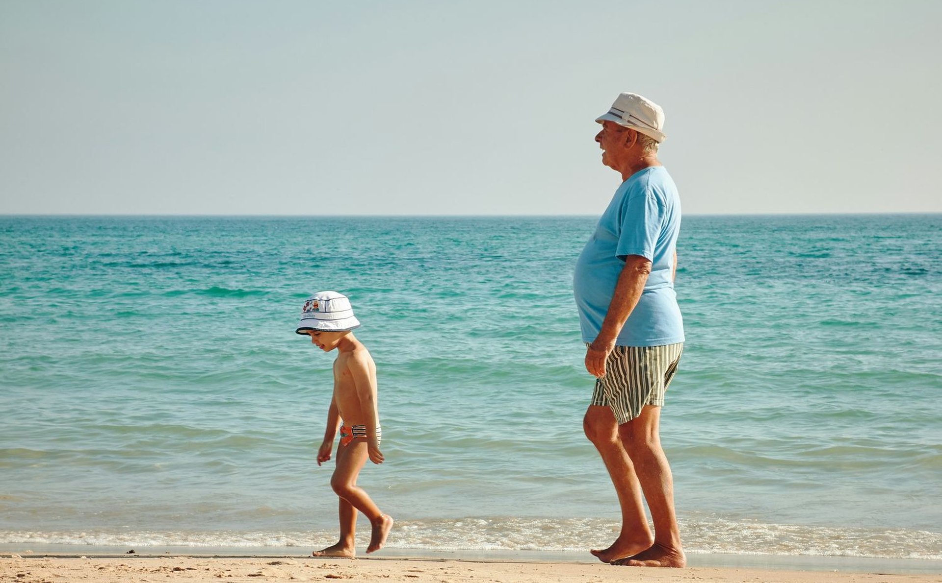 An older man in a sun hat walking along the shoreline with his grandchild